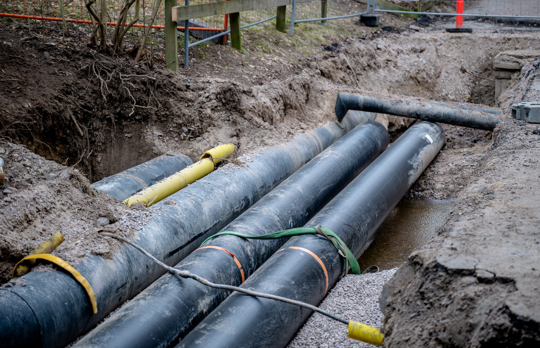 Large black underground pipes being installed in an excavated trench at a construction site, surrounded by dirt and gravel, with safety barriers